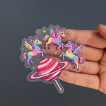 Hand holding a clear sticker with colorful pegasus-like creatures and a striped lollipop on a dark background