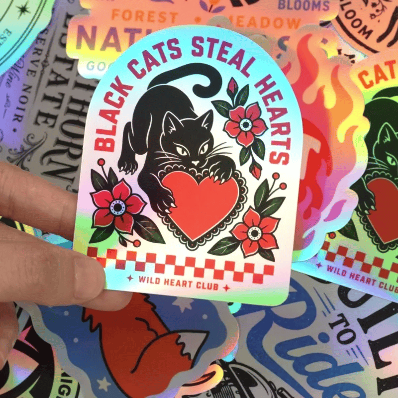 Hand holding a holographic black cat sticker with red heart and floral artwork. Dynamic holographic shine shifts with the light, revealing changing colors from different angles.