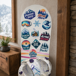 A white snowboard covered in colorful snowboard brand stickers and decals placed indoors near a fireplace.