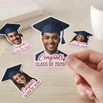 Hand peeling a custom Congrats Class of 2026 photo sticker with a graduate wearing a navy blue cap and gown.