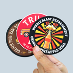 Hand holding three custom circle stickers with colorful soda, truck and honey logo designs.