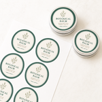 Custom-printed circle vinyl labels for botanical balm applied to silver cosmetic tins, demonstrating clean die-cut edges and elegant brand presentation.