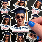 Hand holding a custom Class of 2026 graduation face sticker with a mortarboard hat, surrounded by other personalized grad stickers on a dark surface.