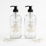 Two large glass pump bottles for hand and dish soap featuring elegant gold-printed clear labels with wave and hand icons.
