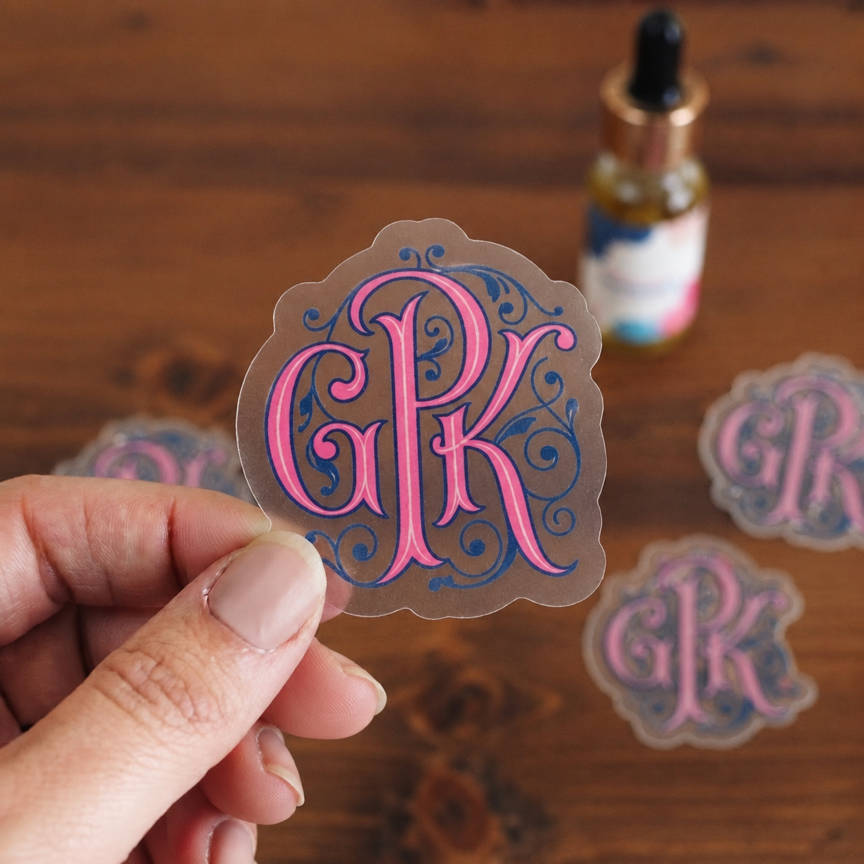 Hand holding a custom die-cut clear monogram sticker with pink and blue lettering.