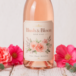 A "Blush & Bloom" custom wine label with watercolor pink rose illustrations, applied to a Rosé bottle and surrounded by fresh flowers.