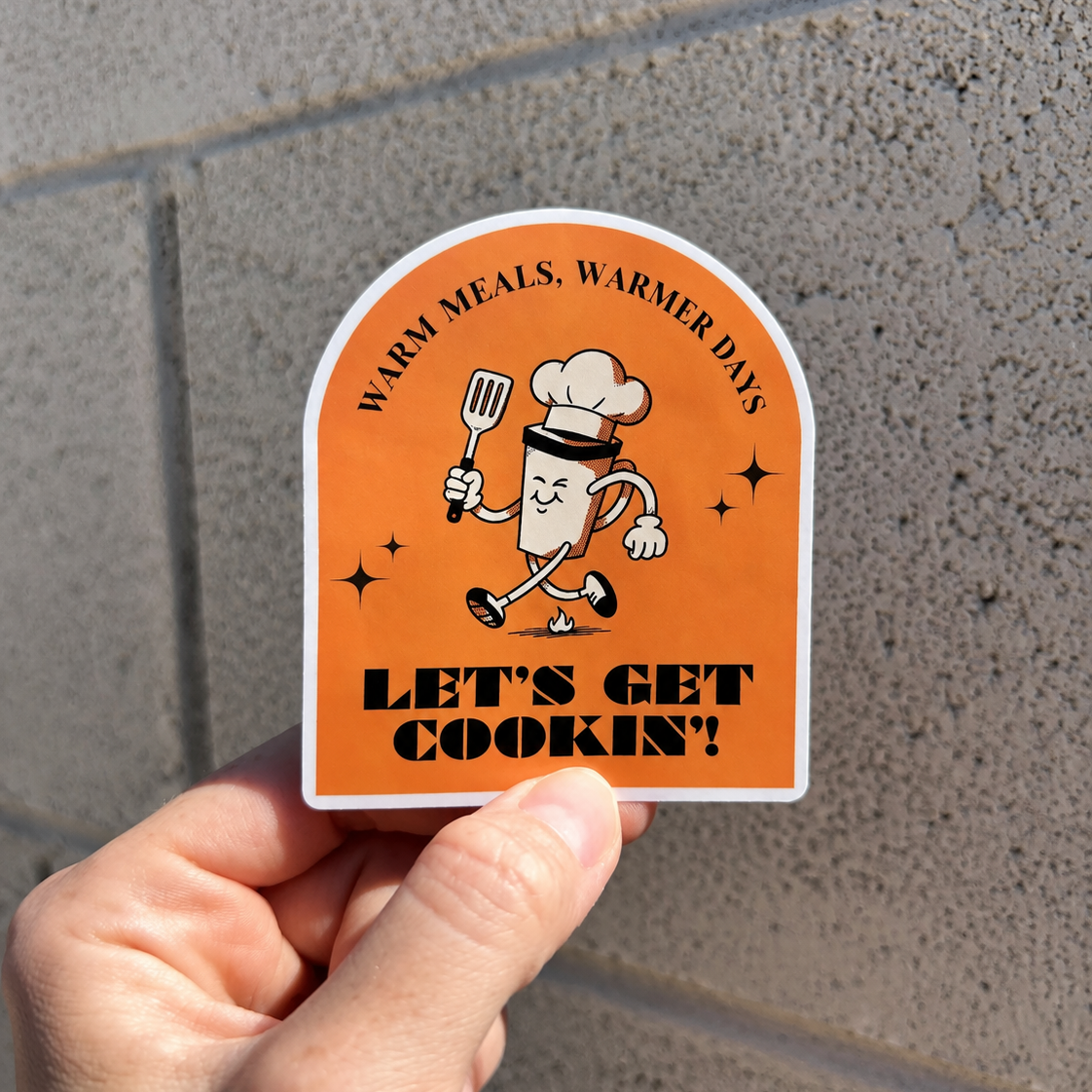 Custom cooking-themed giveaway sticker with bold black text and cartoon chef artwork, held outdoors in natural light.
