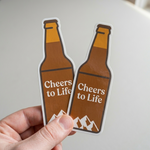 Two custom glossy vinyl stickers shaped like beer bottles with "Cheers to Life" text and mountain graphics.