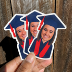 Hand holding custom graduate face stickers with a navy blue cap and a red graduation stole against a wooden background.