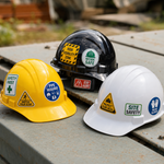 Three construction hard hats in yellow, black, and white colors, featuring safety decals such as Safety First, Wear Gloves, and Watch Your Step.