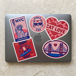 A sleek laptop decorated with cute cat, NYC, and golden gate bridge themed stickers in red, blue, and pink colors.