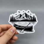 A custom matte vinyl sticker featuring a black and white illustration of a canoe on a lake with mountains and pine trees in the background, held by a hand to show scale.