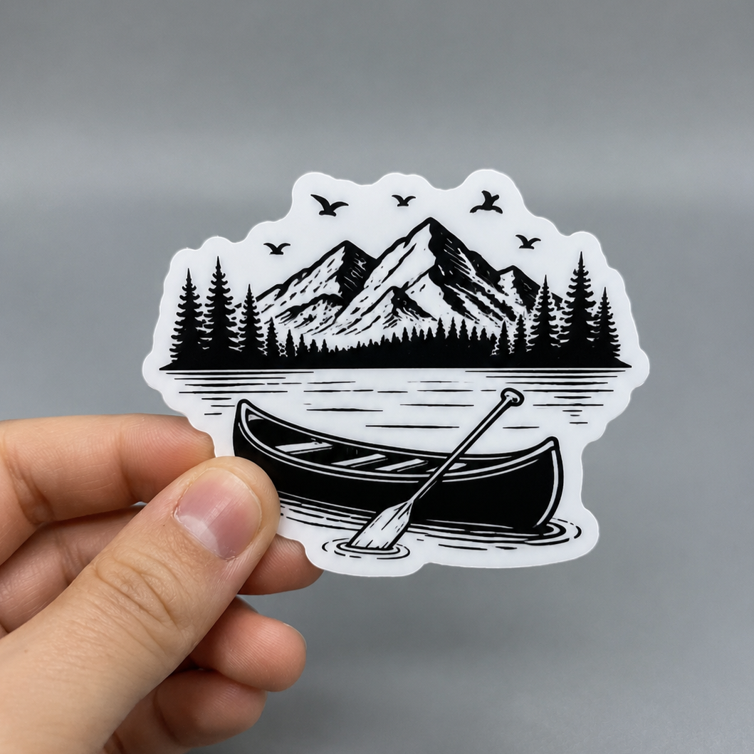 A custom matte vinyl sticker featuring a black and white illustration of a canoe on a lake with mountains and pine trees in the background, held by a hand to show scale.