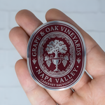 Hand holding a custom oval silver metallic wine logo sticker with a burgundy vineyard design.