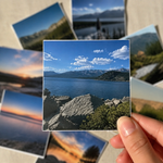 A hand holds a square photo sticker print showing a mountain lake landscape with blue sky and white clouds, while several other landscape photo prints are scattered in the background on a light surface.