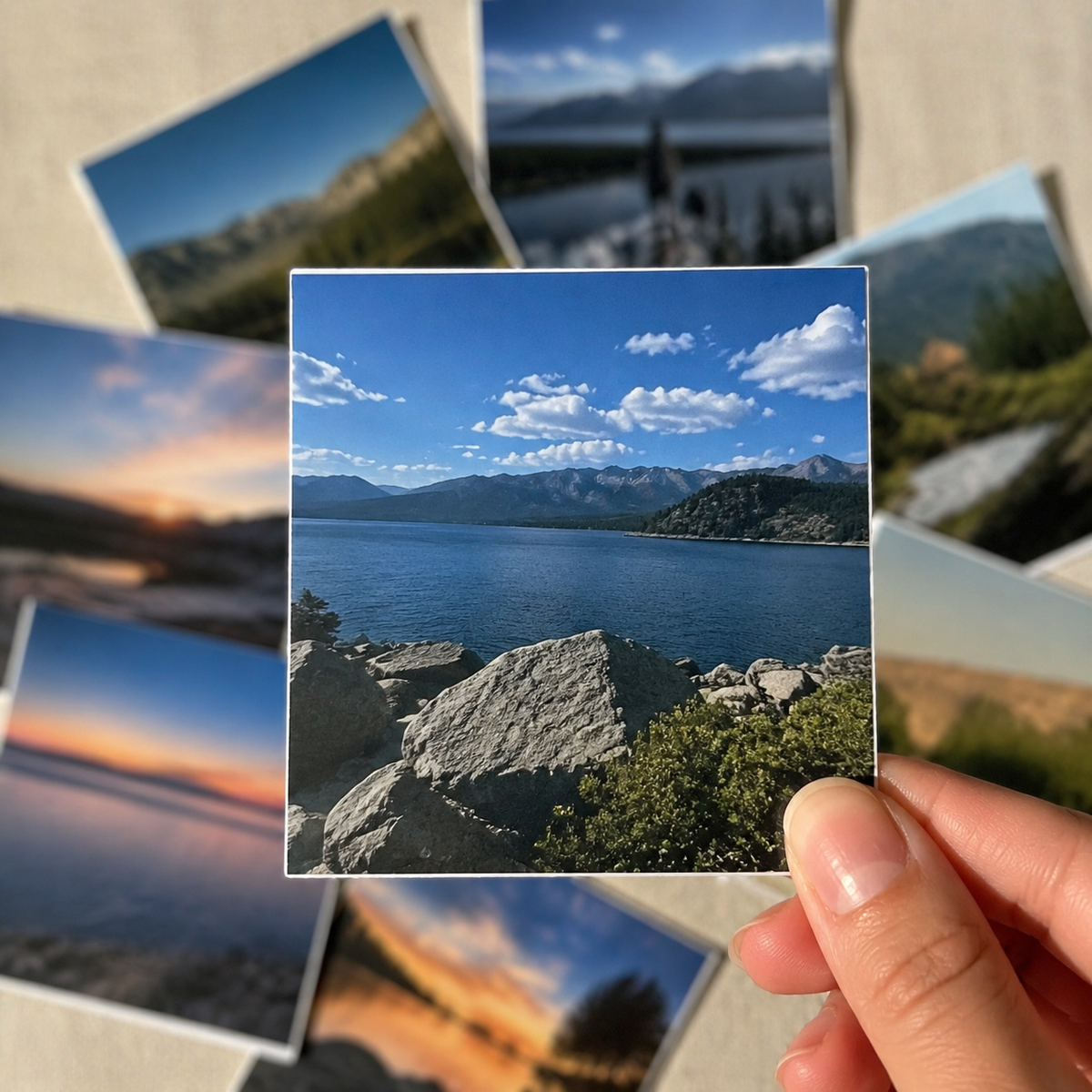 A hand holds a square photo sticker print showing a mountain lake landscape with blue sky and white clouds, while several other landscape photo prints are scattered in the background on a light surface.