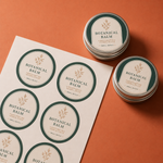 Matte finish custom round vinyl labels on a sheet and applied to aluminum cosmetic tins for a handcrafted botanical balm brand.