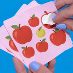 Hand applying sticker stickers of red and green apples design onto pink backing paper against a blue background