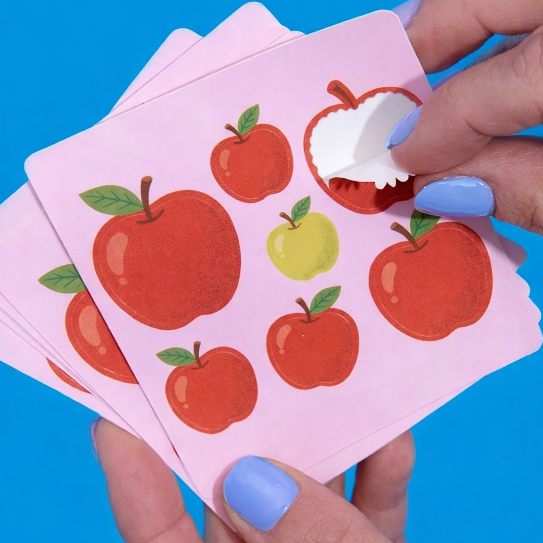 Hand applying sticker stickers of red and green apples design onto pink backing paper against a blue background