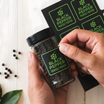 Hands applying a custom vinyl black pepper label to a clear spice jar beside a sheet of matching labels.