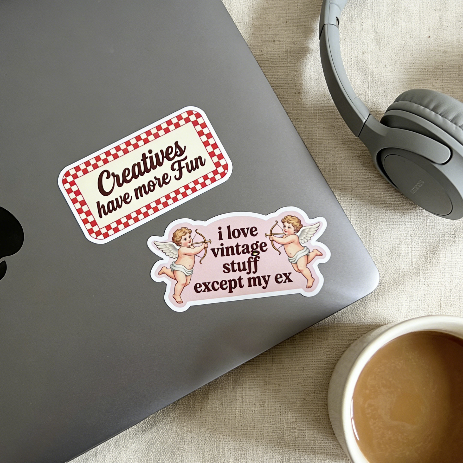 Two high-definition custom vinyl stickers placed on a silver laptop next to headphones and a coffee cup, featuring a red checkered 'Creatives have more Fun' sticker and a vintage cupid 'i love vintage stuff except my ex' sticker.