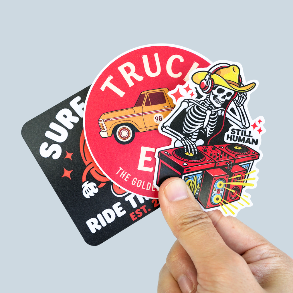 Hand holding colorful custom vinyl stickers featuring truck, surf and skeleton DJ designs.