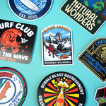 Hand holding colorful custom vinyl stickers featuring truck, surf and skeleton DJ designs.