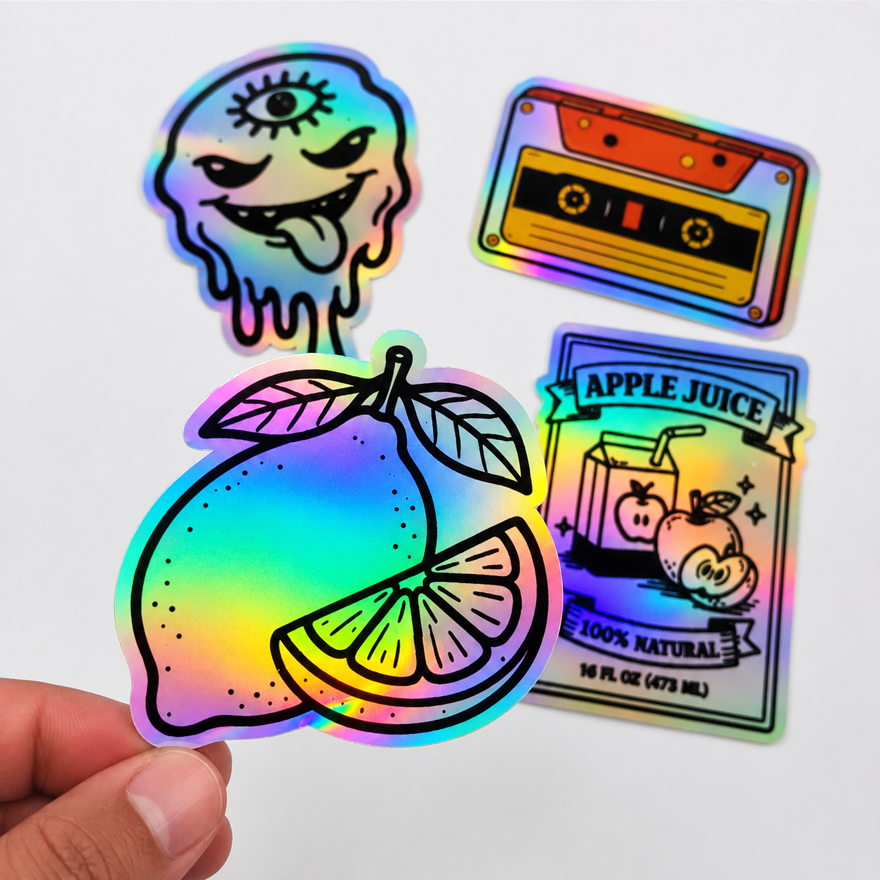A hand holding a custom holographic lemon sticker with a vibrant rainbow shimmer effect, displayed alongside other holographic decals including a cassette tape and a juice carton.