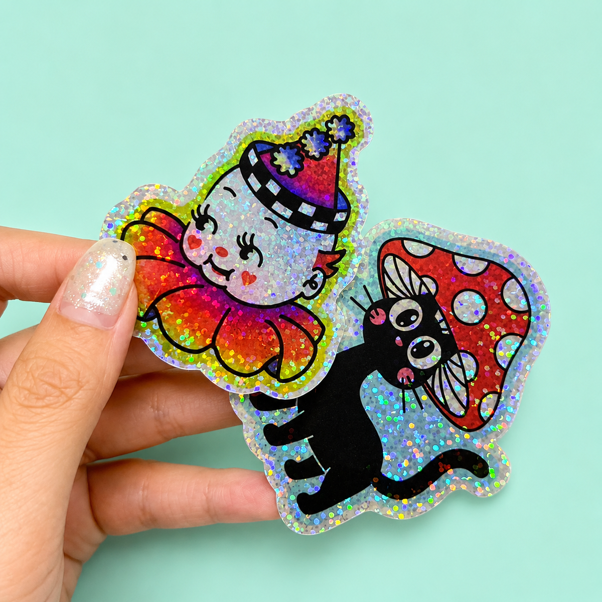 Hand holding two colorful glitter stickers featuring a party clown and a cat under a mushroom.