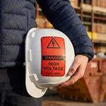 A close-up shot of a construction worker holding a white hard hat. A red and black heavy-duty danger label is pasted on the side of the helmet