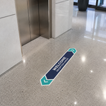 A long, arrow-shaped floor decal in a modern office lobby near elevators, featuring the text "WELCOME - RECEPTION AHEAD" in navy blue and teal.