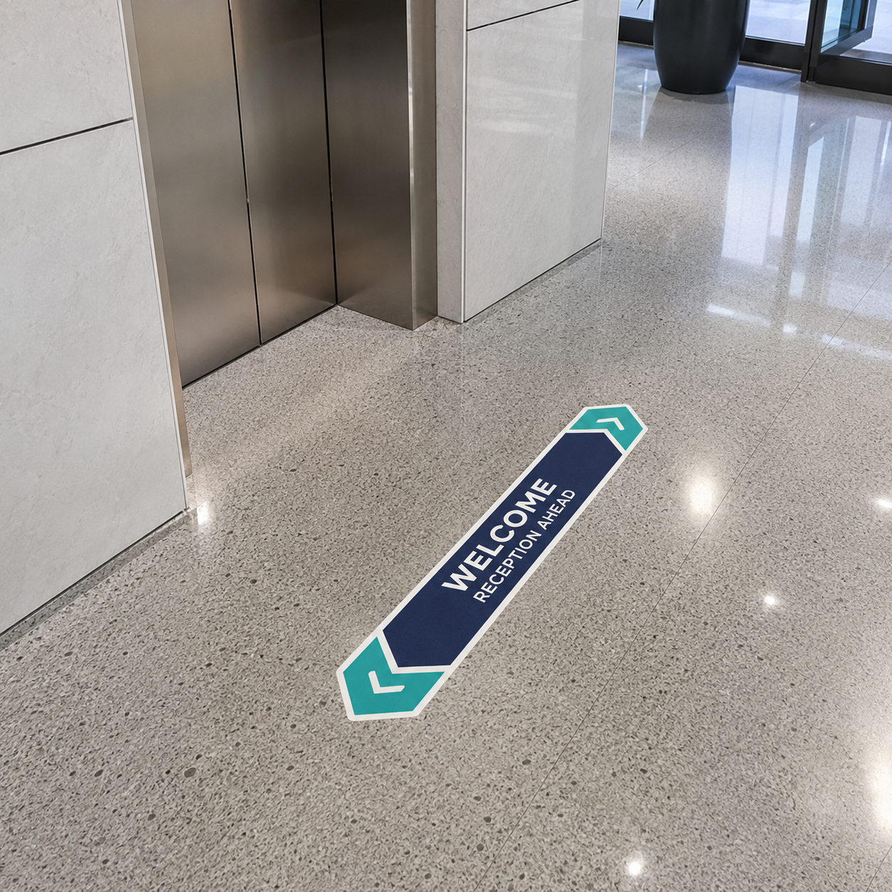 A long, arrow-shaped floor decal in a modern office lobby near elevators, featuring the text "WELCOME - RECEPTION AHEAD" in navy blue and teal.