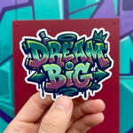 A vibrant die-cut sticker featuring "Dream Big" in complex graffiti lettering with green, purple, and yellow gradients, held in front of an urban mural.