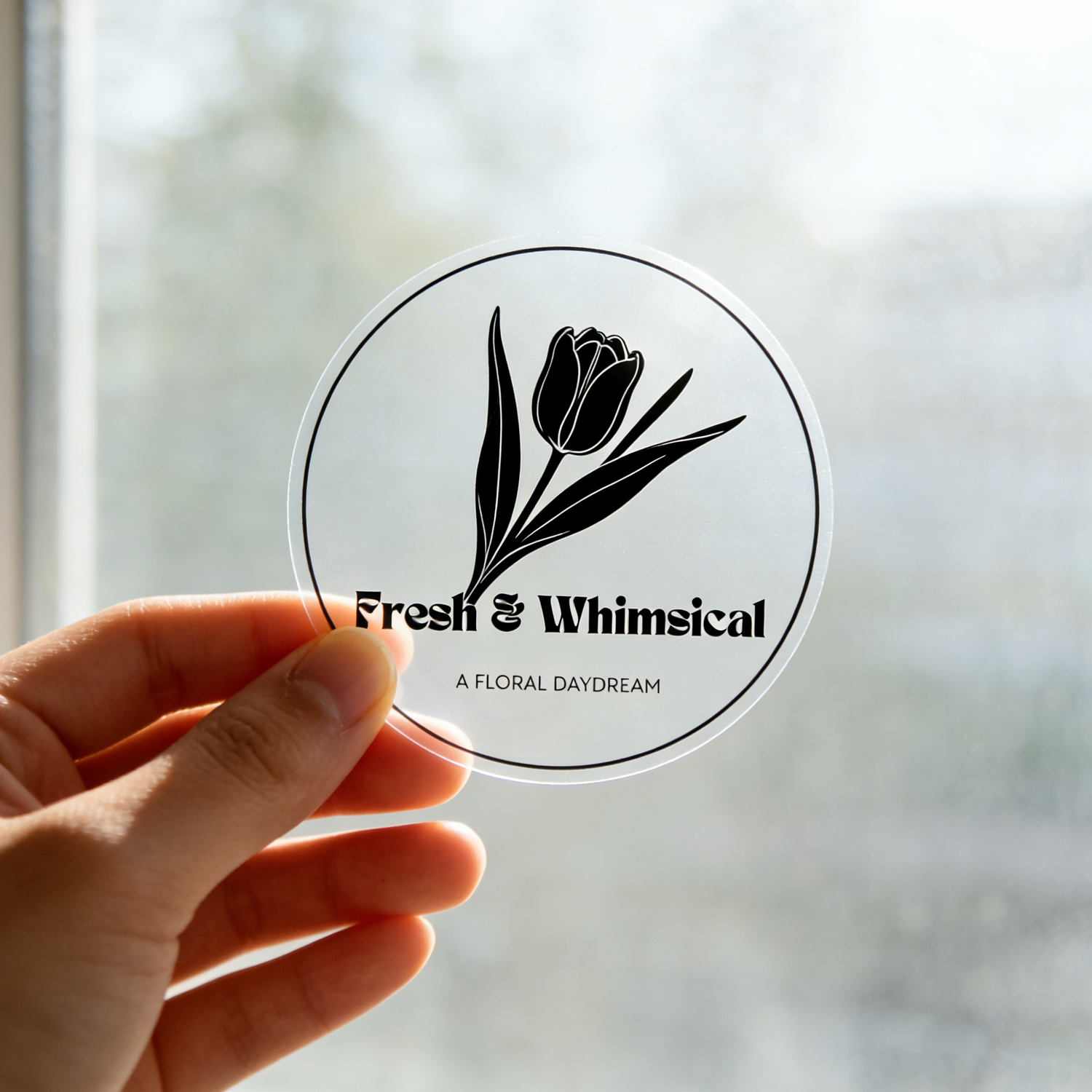 Hand holding a round clear vinyl sticker with a tulip design and text against a blurred window background