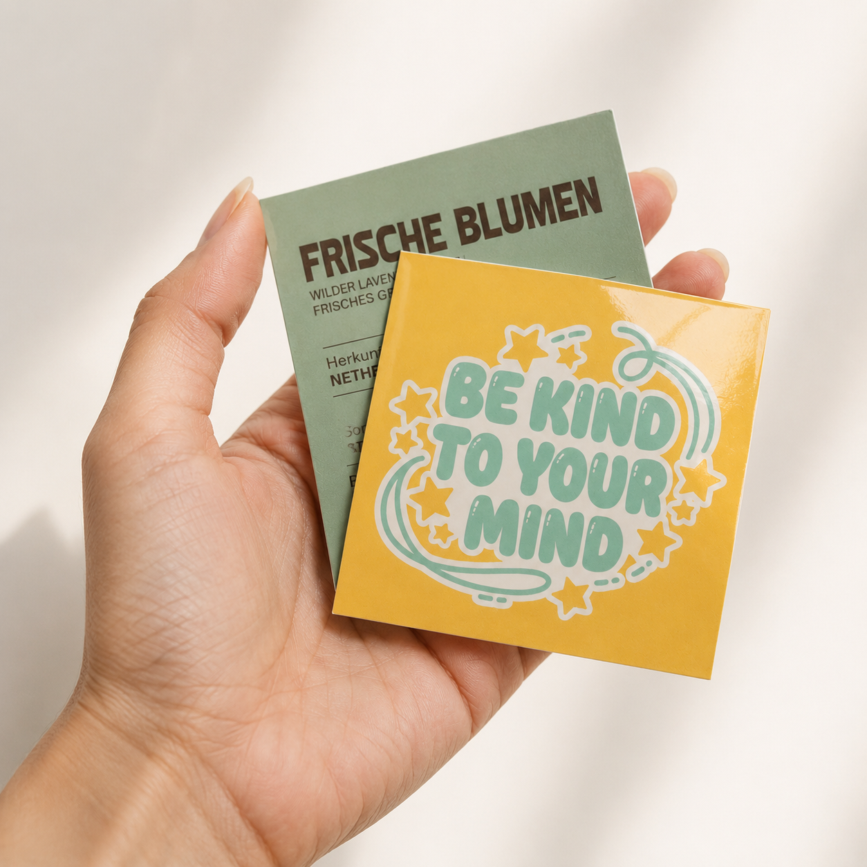 Hand holding glossy custom square stickers with a Be Kind design