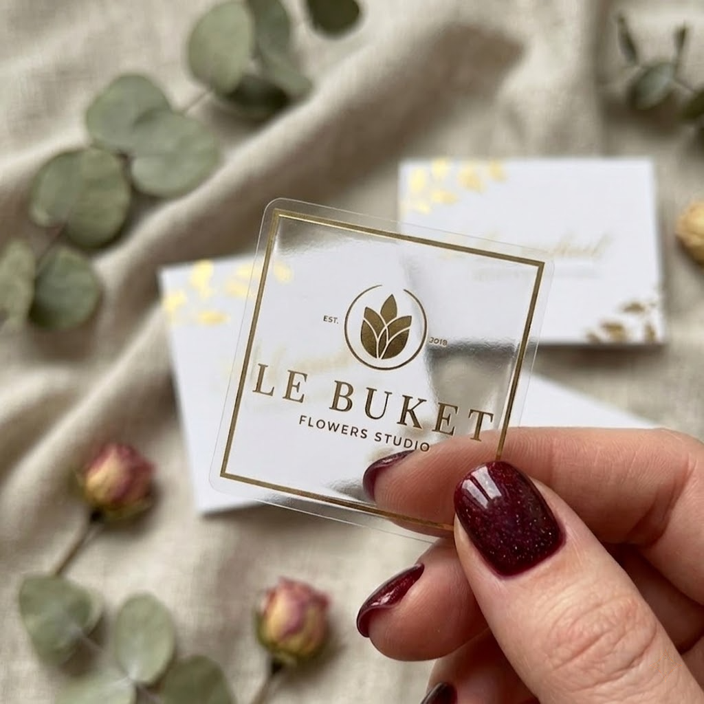A close-up of a premium square clear label with a gold foil logo for "LE BUKET Flowers Studio" held against a soft fabric background.