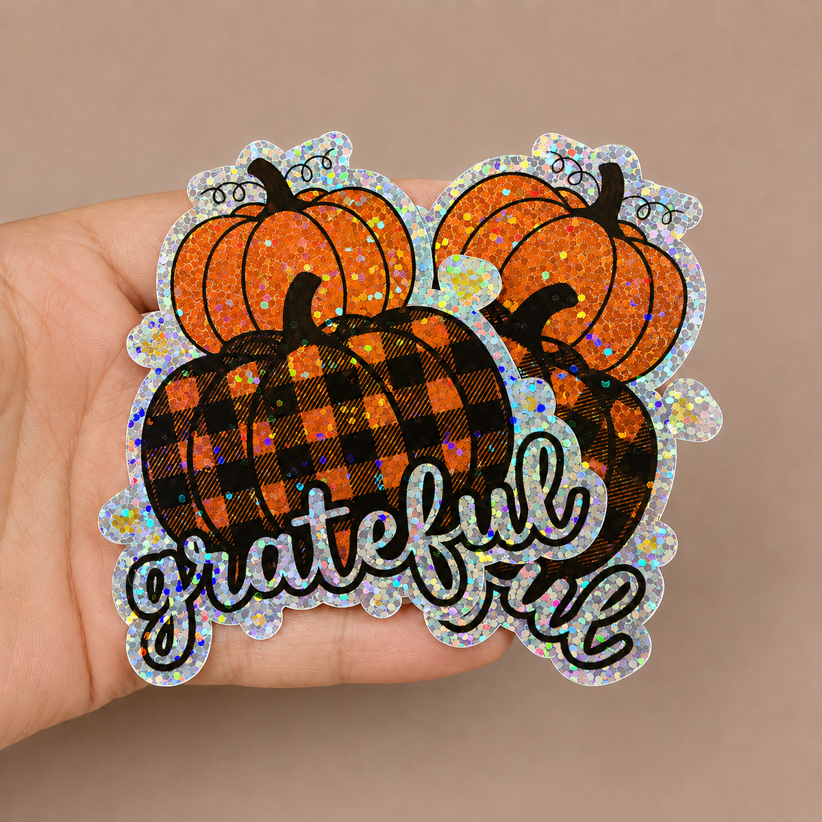 Hand holding layered pumpkin glitter stickers with a black plaid design and grateful script lettering.