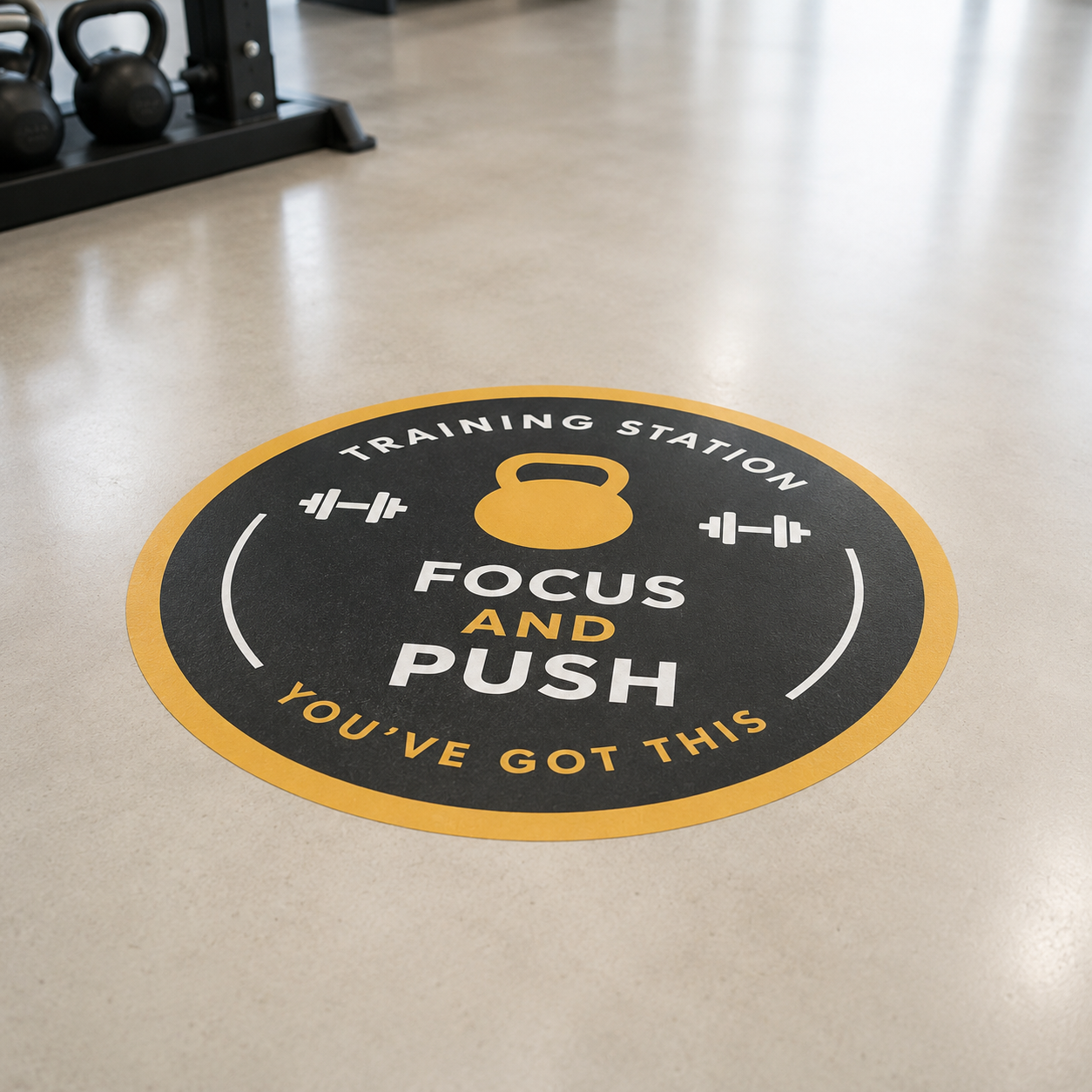 A circular black and yellow floor decal in a gym setting that reads "TRAINING STATION - FOCUS AND PUSH - YOU'VE GOT THIS" with a kettlebell icon.