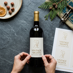 Hands carefully applying a "Vintage Nights" custom vinyl wine sticker to a dark bottle, demonstrating the ease of application for DIY gift projects.