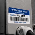 A close-up of a permanent blue and white industrial asset sticker labeled Operational Asset with an ID EQ-3487 and a barcode, applied to gray metal machinery with water droplets.