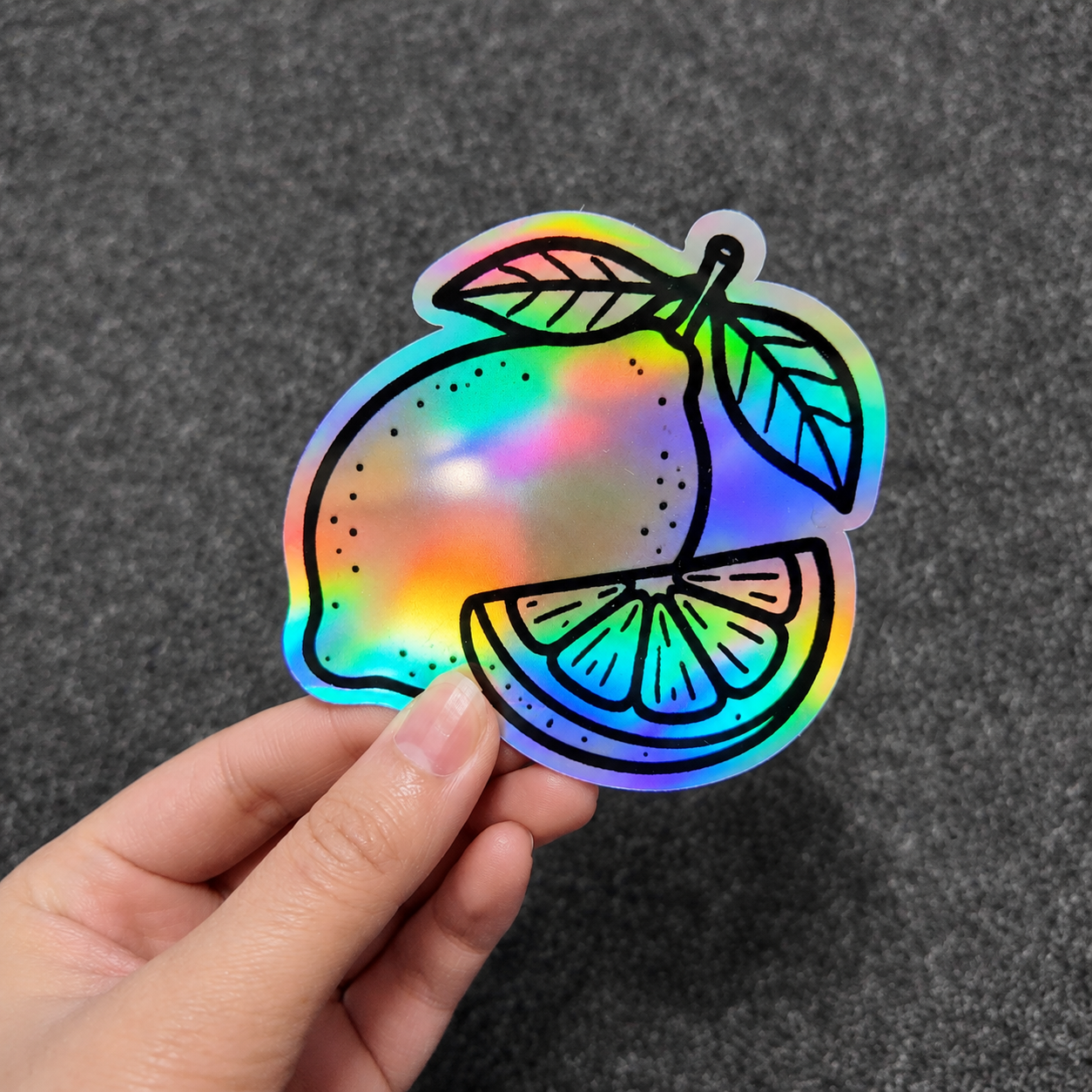 A hand holding a holographic lemon sticker made of premium vinyl, showcasing a vibrant rainbow iridescent finish and durable industrial-grade quality against a dark textured background.