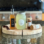 A close-up shot on a natural wood serving board on a kitchen counter, featuring glass jars filled with fresh cucumber juice, artisanal ginger tea, cinnamon sticks, and a small jar of coarse sea salt