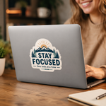 Person using a laptop with a 'Stay Focused' vinyl decal on a wooden desk.