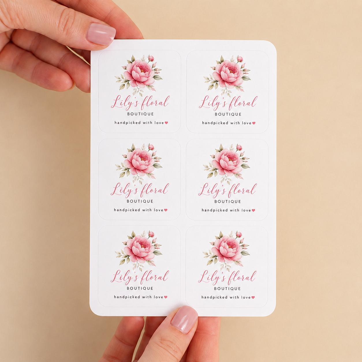 Sheet of six "Lily's Floral Boutique" custom labels featuring pink roses and green leaves, with the text "handpicked with love" at the bottom, held by hands against a beige background.