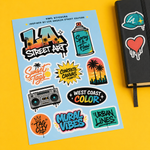 Top-down premium product photography of an LA street art vinyl sticker sheet with vibrant graffiti designs, a boombox, and spray cans on a bright yellow backdrop next to a black notebook.