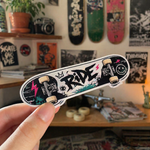 Hand holding a miniature skateboard model with skateboarding stickers, in front of a background of customized skateboards.