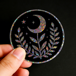Hand holding a glitter sticker with designs of  moon, star, and leaf design on a black background