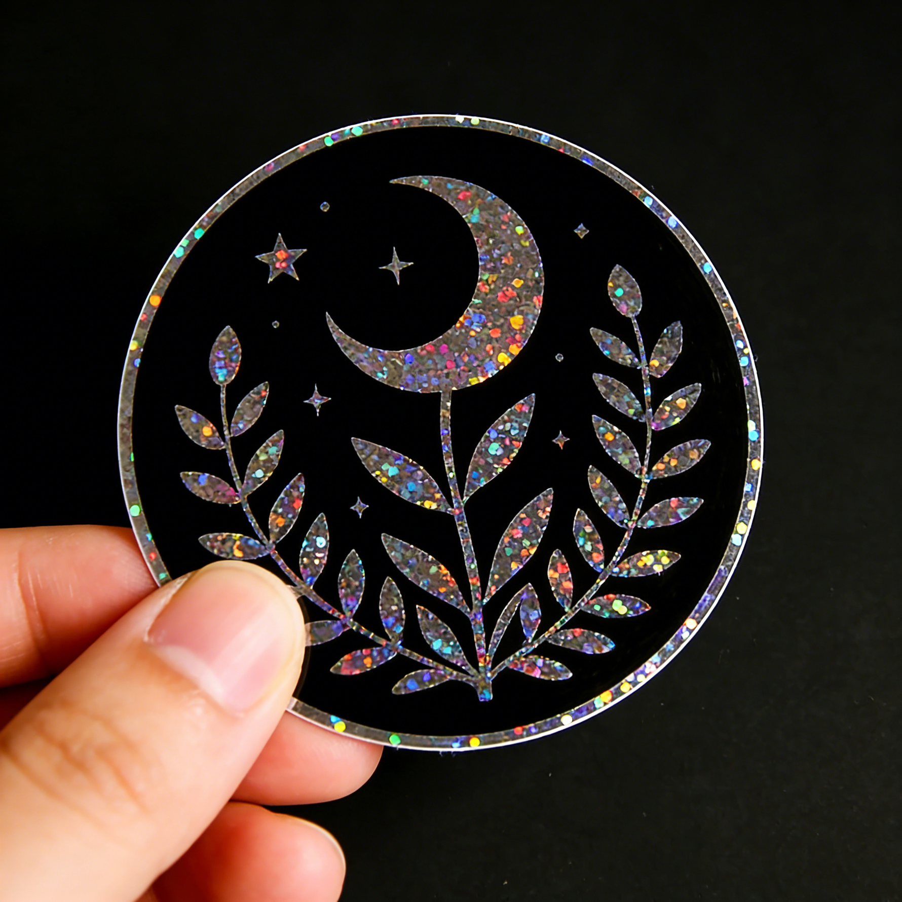 Hand holding a glitter sticker with designs of  moon, star, and leaf design on a black background