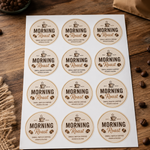 Top-down view of a printed sheet of 12 circular coffee brand stickers with a vintage aesthetic, featuring 'Morning Roast' logo on a wooden background.