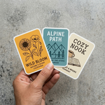 Three nature themed rounded corner stickers held by a hand on a concrete surface, waterproof and UV-resistant